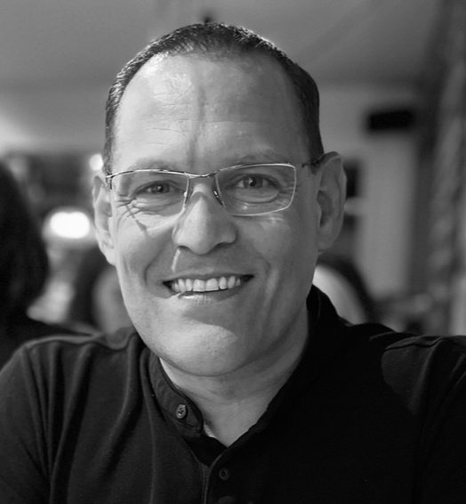 Ian Ashby Smiling man wearing glasses, seated in a casual setting with a blurred background.